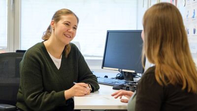 En kvinnelig fastlege i grønn genser sitter ved et skrivebord og smiler mens hun lytter til en annen kvinne som sitter overfor henne. De ser ut til å være i en samtale på et kontor. På bordet ligger et ark og en penn, og en datamaskin står i bakgrunnen.