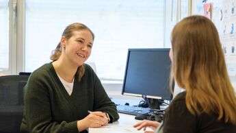 En kvinnelig fastlege i grønn genser sitter ved et skrivebord og smiler mens hun lytter til en annen kvinne som sitter overfor henne. De ser ut til å være i en samtale på et kontor. På bordet ligger et ark og en penn, og en datamaskin står i bakgrunnen.