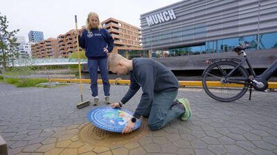 En mann fester et emblem til et kumlokk. En dame står bak med en feiekost.