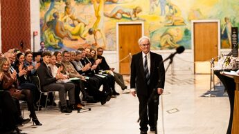 Prisvinner går frem mot en et podium og en mikrofon for å ta i mot en pris. Publikum sitter med ansiktet vendt mot prisvinner og klapper.