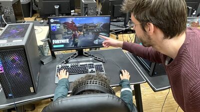 Mann peker på pc-skjerm der det spilles Fortnite. Bakhodet på en ungdom synes.
