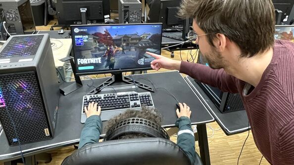 Mann peker på pc-skjerm der det spilles Fortnite. Bakhodet på en ungdom synes.