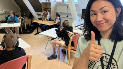 Dame viser tommelen opp med elever i bakgrunnen. Elevene lager musikk på laptop.