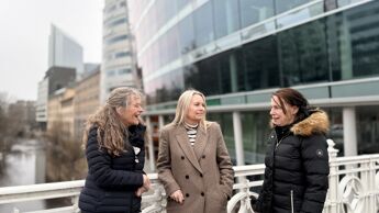 Tre kvinner på en bro, med storbybygninger og elv bak.