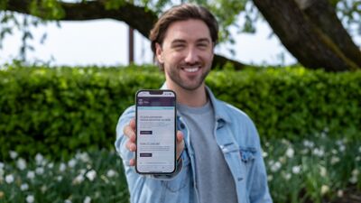 Ung mann viser fram en mobilskjerm med nyhetsbrev fra Oslo kommune. Grønne busker bak han.