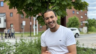 Smilende ung mann i hvit t-skjorte med hestehale foran tre.