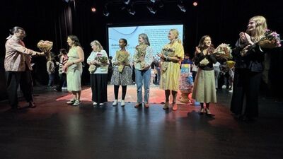 8 mennesker i halvsirkel. Klaapper og mottar blomster. Inne på scene.