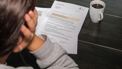 Sittende kvinne bakfra med brev fra det offentlige på borde og en kaffekopp foran seg.