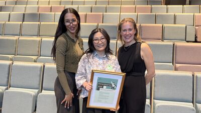 Tre kvinner i tomt auditorium smiler. Kvinne i midten holder opp diplom.