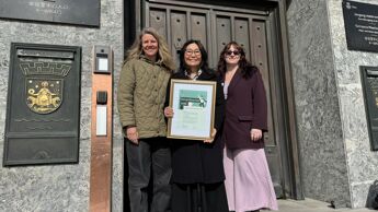 Tre kvinner utenfor bygning. En holder innrammet diplom.