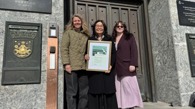 Tre kvinner utenfor bygning. En holder innrammet diplom.