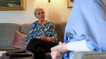 Bilde av eldre dame i sofaen hjemme hos seg selv med deler av en hjemmesykepleier i blått arbeidstøy i forgrunnen