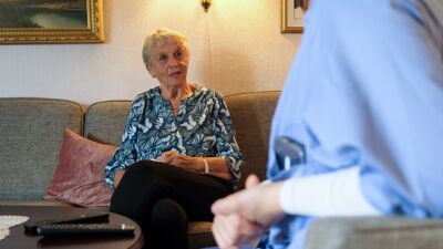 Bilde av eldre dame i sofaen hjemme hos seg selv med deler av en hjemmesykepleier i blått arbeidstøy i forgrunnen