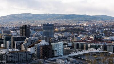 Oversiktsbilde av Oslo sentrum med moderne bygninger i Barcode-rekken, den hvite broen Akrobaten, og et bakteppe av skogkledde åser. En blanding av urbane og naturlige elementer preger utsikten.