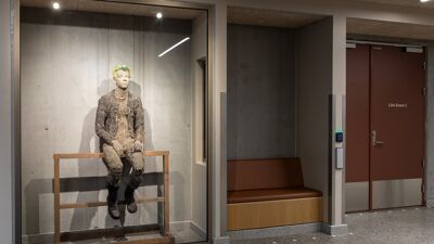 Interiørfoto av en korridor i et offentlig bygg. I en glassmonter sitter en figurativ skulptur av en person på en enkel trekonstruksjon. Skulpturen har grove, jordfargede overflater og sitter med bena hengende ned. Rundt monteren er det betongvegger, benk og dører til rom i bygget.