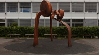 Fargefotografi av en stor, abstrakt skulptur i rustfarget metall plassert utendørs. Skulpturen består av buede og sirkulære former som står på tre ben. Den er plassert på et rundt underlag, med benker og grønt hekkeparti rundt. I bakgrunnen ses fasaden på en skolebygning med vinduer og persienner.