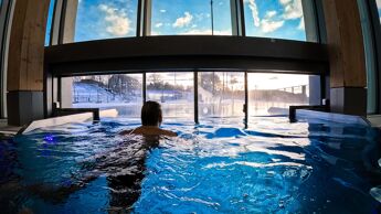 En person som svømmer i et oppvarmet basseng på det nye Tøyenbadet, med utsikt mot vinterlandskap og blå himmel gjennom store vinduer.