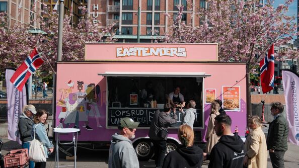 Rosa foodtruck foran The Hub. Person i trucken serverer en "loaded fries". Folk foran trucken. Det er sol. Trucken har norske flagg.