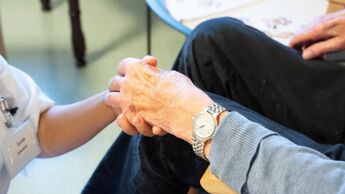 Sykepleier holder hånden til en eldre menneske som sitter i en stol.