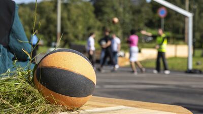 Fokus på en basketball som ligger på en benk ute på en basketball bane der det står flere i bakgrunn som spiller.