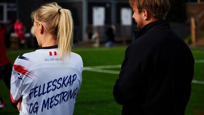 To personer som ser fotball. Den ene har drakt med teksten "fellesskap og mestring"