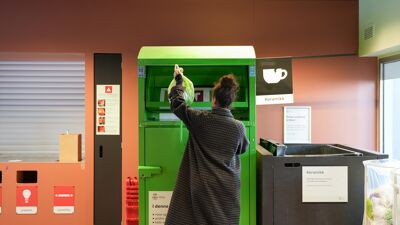 Dame med ryggen til løfter en grønn plastpose foran en grønn container. Veggen bak er rød. Det er en gjenvinningsstasjon.