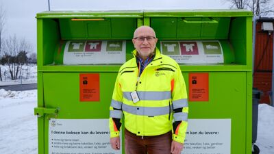 Mann i gul refleksjakke foran to grønne klescontainere. Det er snø på bakken.
