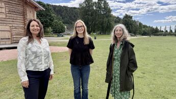 Tre personer står på en gresslette i et parkområde ved en trebygning.