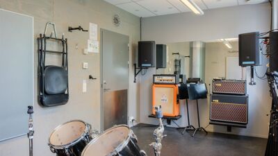 Innendørs musikkøvingsrom med trommesett i forgrunnen. Langs veggene står gitarforsterkere, høyttalere og notestativer, og en stor veggspeil dekker deler av bakveggen.