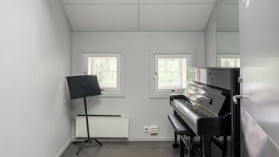 Øvingsrom med oppreist piano, pianokrakk og notestativ. Rommet har to vinduer og lyse vegger.