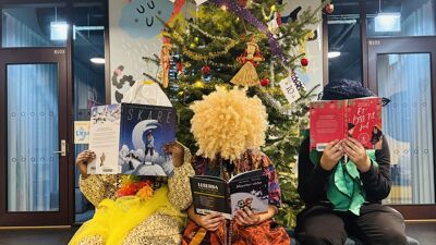 Tre mennesker sitter utkledd foran et juleferie. De dekker ansiktet med en bok.