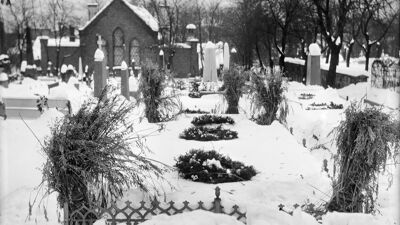 Snødekt kirkegård med gravsteiner, kranser og beplantning rundt gravene. I bakgrunnen står en liten murbygning omgitt av nakne trær.
