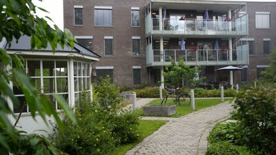 Bilde av mursteinsbygg med viduer og veranfaer i tre etasjer, en gangvei og et lite hagehus med vinduer i front og grønt gress og vegetasjon (busker og trær)