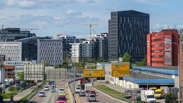 Motorvei med biler som kjører i begge retninger. Skilt henger over veien. Høye bygninger i horisonten.