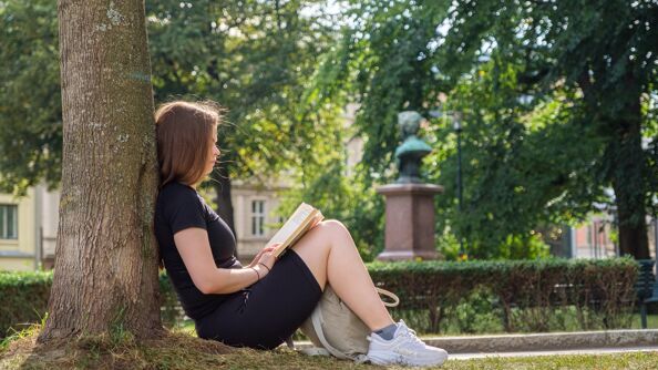 En person sitter med ryggen inntil et tre og leer i en bok i en park.