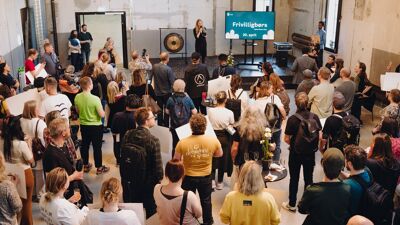 Bilde av mange mennesker i et rom foran en scene med konferansier