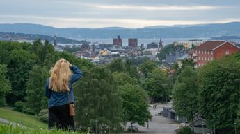En kvinne med langt lyst hår, kledd i blå jeansjakke (olajakke), svarte bukser og med en liten brun skulderveske, står med ryggen til på en grønn gressplen i Torshovdalen og ser ut over byen. I mellomgrunnen er det et parkområde med gangstier og trær. I bakgrunnen ses Oslo sentrum med landemerker som Oslo rådhus, Oslofjorden og åser i det fjerne.