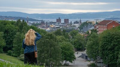 En kvinne med langt lyst hår, kledd i blå jeansjakke (olajakke), svarte bukser og med en liten brun skulderveske, står med ryggen til på en grønn gressplen i Torshovdalen og ser ut over byen. I mellomgrunnen er det et parkområde med gangstier og trær. I bakgrunnen ses Oslo sentrum med landemerker som Oslo rådhus, Oslofjorden og åser i det fjerne.
