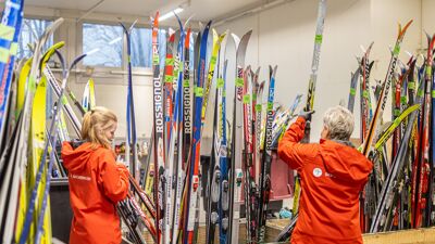 To kvinner i røde jakker foran store kasser med ski.