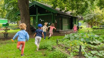 Fire barnehagebarn som løper etter hverandre i en park med trær og planter rundt seg