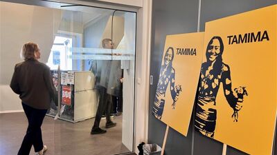 Bilde av to plakater med påskriften TAMIMA og et stensilert bilde av en jente som holder en bunt med blomster, i en kontorlokale. I bakgrunnen går det en mann og en dame.