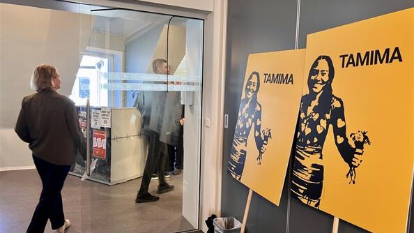 Bilde av to plakater med påskriften TAMIMA og et stensilert bilde av en jente som holder en bunt med blomster, i en kontorlokale. I bakgrunnen går det en mann og en dame.