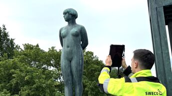 Person i refleksjakke bruker nettbrett til å dokumentere en bronsefigur på sokkel utendørs.