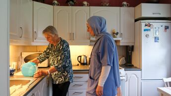 Bilde fra kjøkken hjemme hos en eldre dame som står og ser på en medesindispenser på kjøkkenbenken sammen med en dame fra hjemmetjenesten kledd i arbeidsklær
