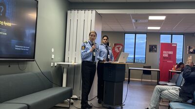 To politibetjenter står foran en folkemengde og presenterer. En person tar bilde med mobilen.