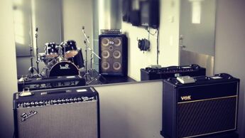 Øvingsrom med trommesett og flere forsterkere plassert foran et stort speil.