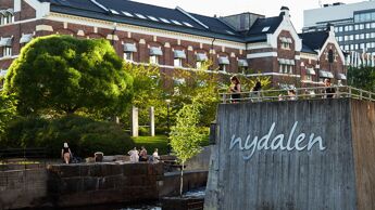En flik av Akerselva og navnet Nydalen festet på en mur som går ut i elva. Teglsteinsbygg i Nylandsveien 21 i bakgrunnen.