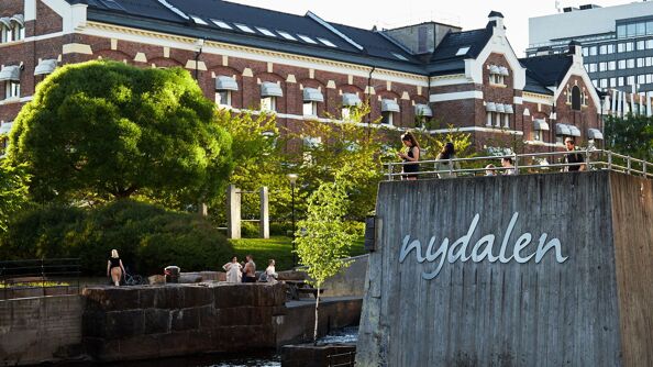 En flik av Akerselva og navnet Nydalen festet på en mur som går ut i elva. Teglsteinsbygg i Nylandsveien 21 i bakgrunnen.