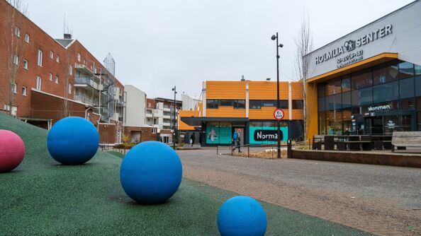 Tre blå og en rosa fastmonterte gummikuler på på grønn gummiplen mellom boliger og senterinngangen på Holmlia.