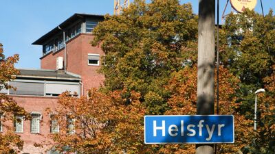 Blått veiskilt som markerer at du har kommet til Helsfyr. Eldre teglsteinsbygg i bakgrunnen.